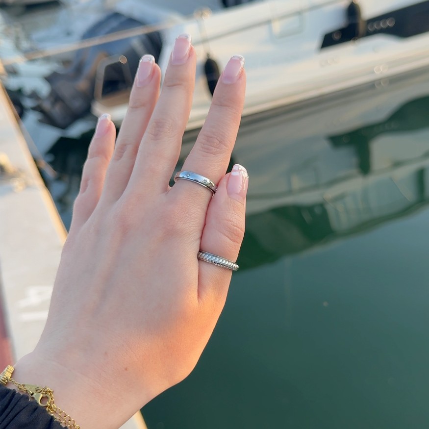 A close-up of a hand with a French manicure and two silver rings, over water with a boat behind.