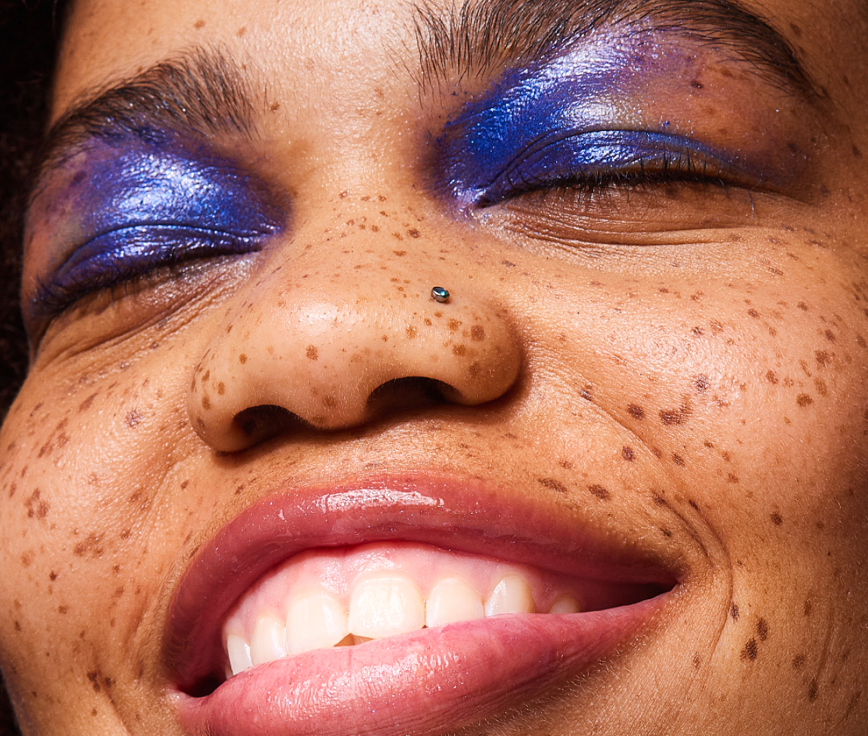A close-up of a smiling person with freckles and shimmery blue eyeshadow, with their eyes closed.