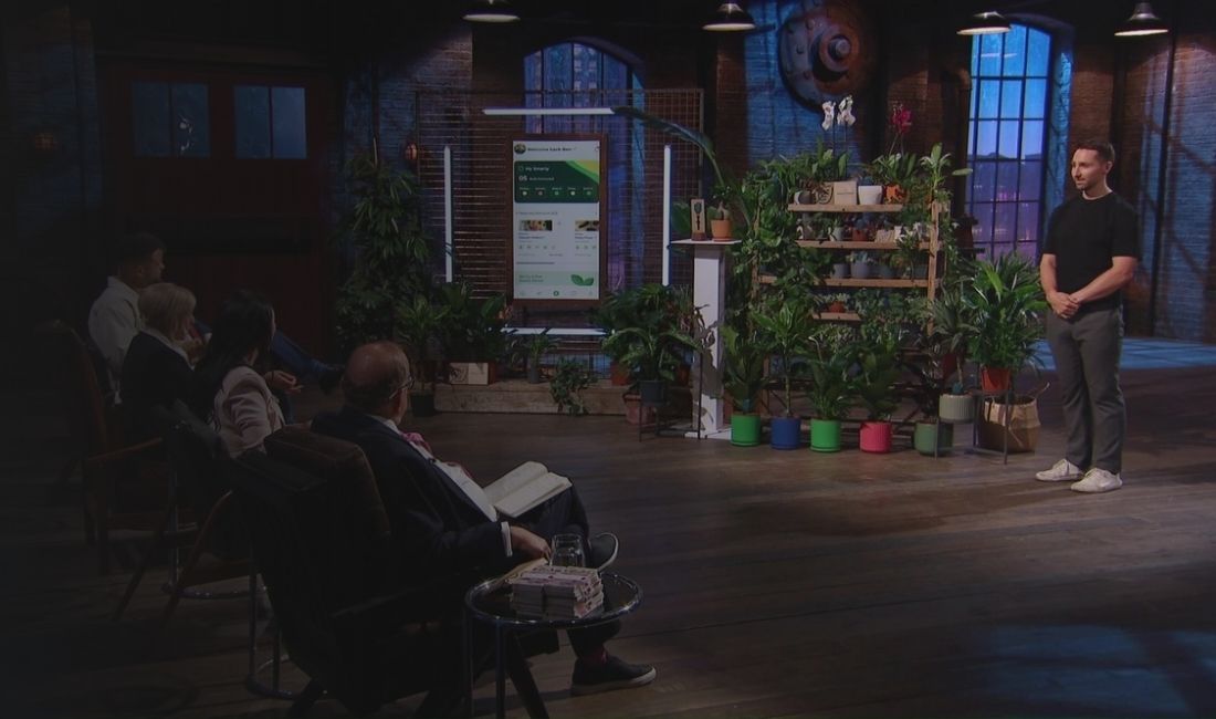 A man presents a plant-related product to a panel of people in a dimly lit studio.