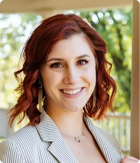 A smiling woman with wavy, red hair, wearing a striped blazer, long earrings, and a necklace.