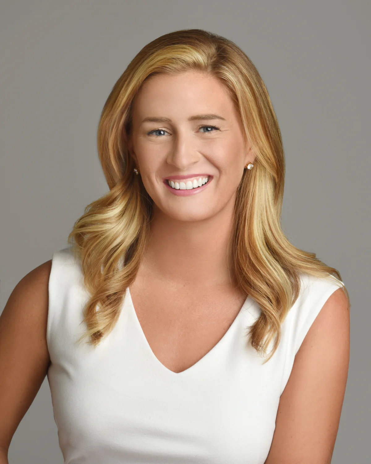 A headshot of a smiling woman with wavy blonde hair wearing a white top against a gray background.
