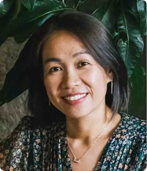 A headshot of a smiling woman with shoulder-length dark hair in front of a large green plant.