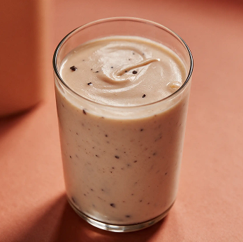 A glass of a creamy, light-colored smoothie with small dark flecks on a pink surface.