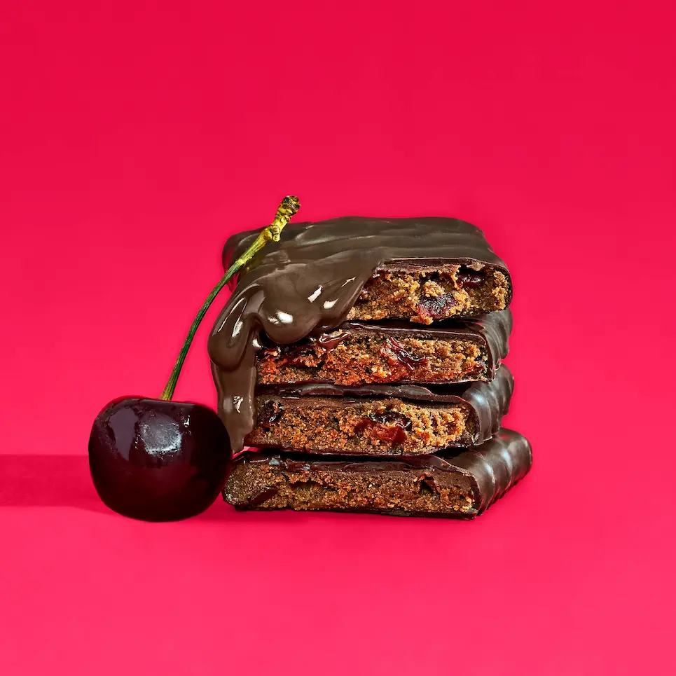 A stack of chocolate-covered cherry bars with chocolate dripping down, next to a fresh cherry, on a pink background.