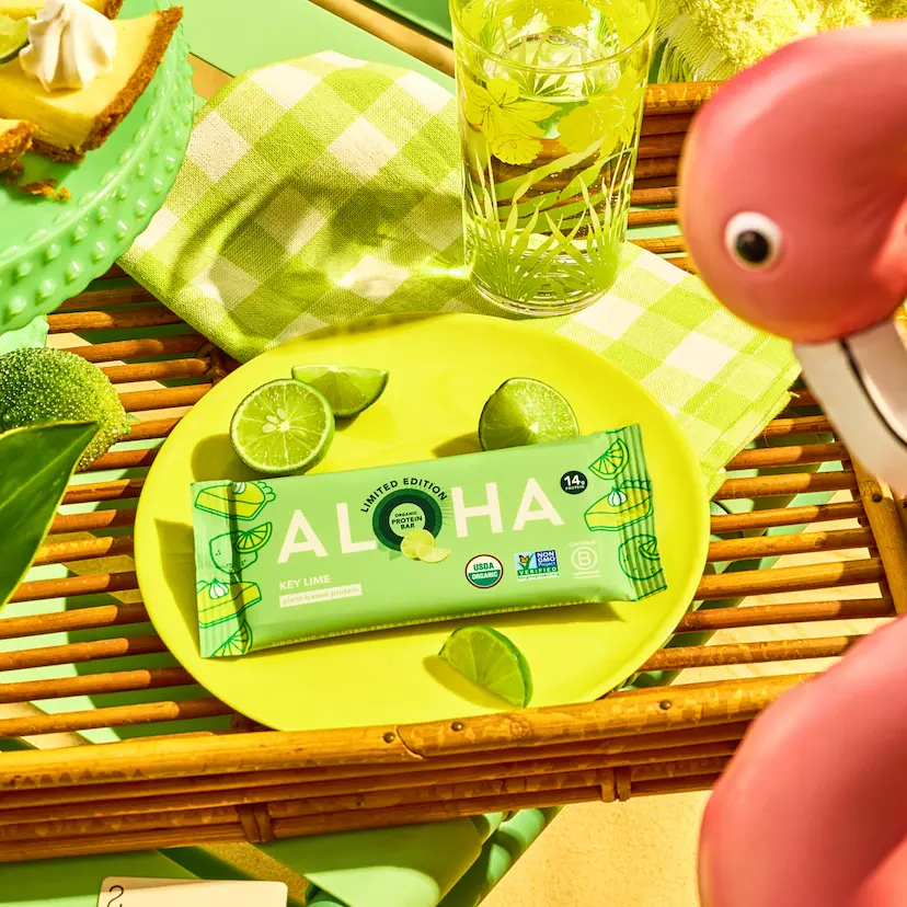 An Aloha Key Lime protein bar on a yellow plate with fresh limes, pie, and a pink flamingo.