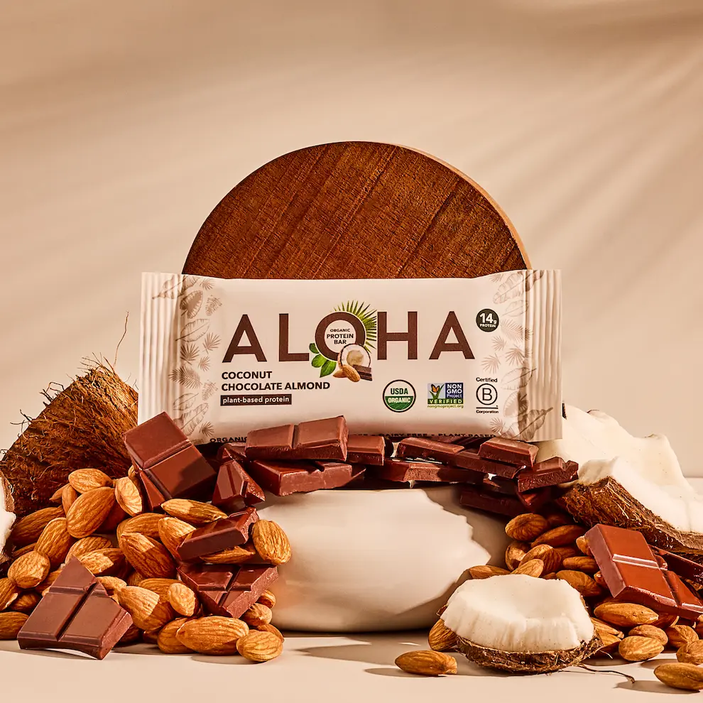 An ALOHA Coconut Chocolate Almond protein bar surrounded by pieces of chocolate, almonds, and fresh coconut.