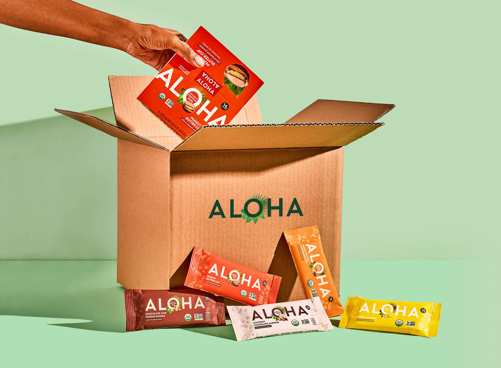 Hand placing Aloha protein bars into a cardboard box on a green background.