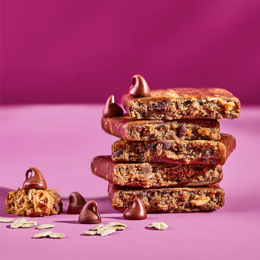 A stack of chocolate chip oat bars with loose chocolate chips and oats against a pink background.