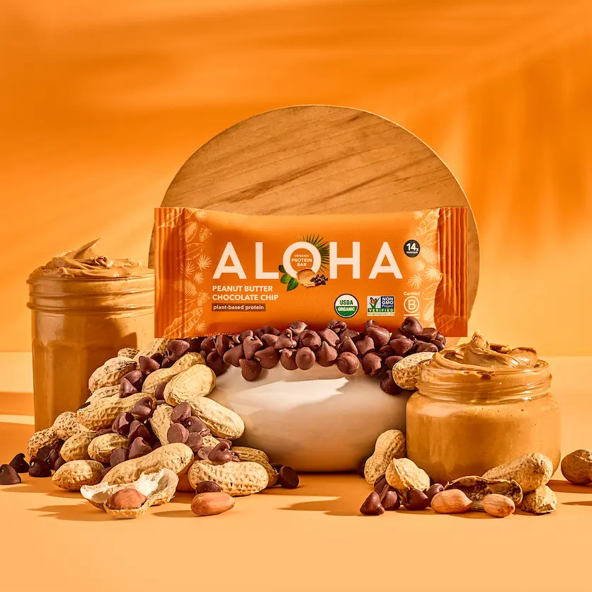 An Aloha Peanut Butter Chocolate Chip protein bar rests among peanuts, chocolate chips, and jars of peanut butter.