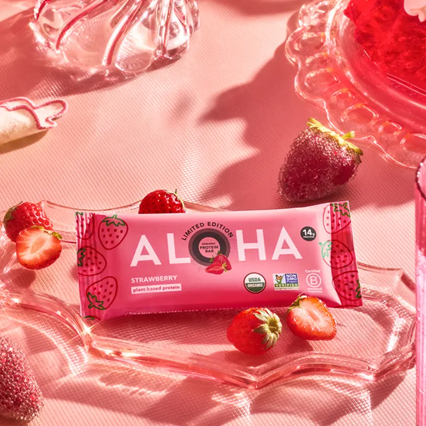 An Aloha strawberry protein bar sits on a decorative pink glass dish surrounded by fresh strawberries.