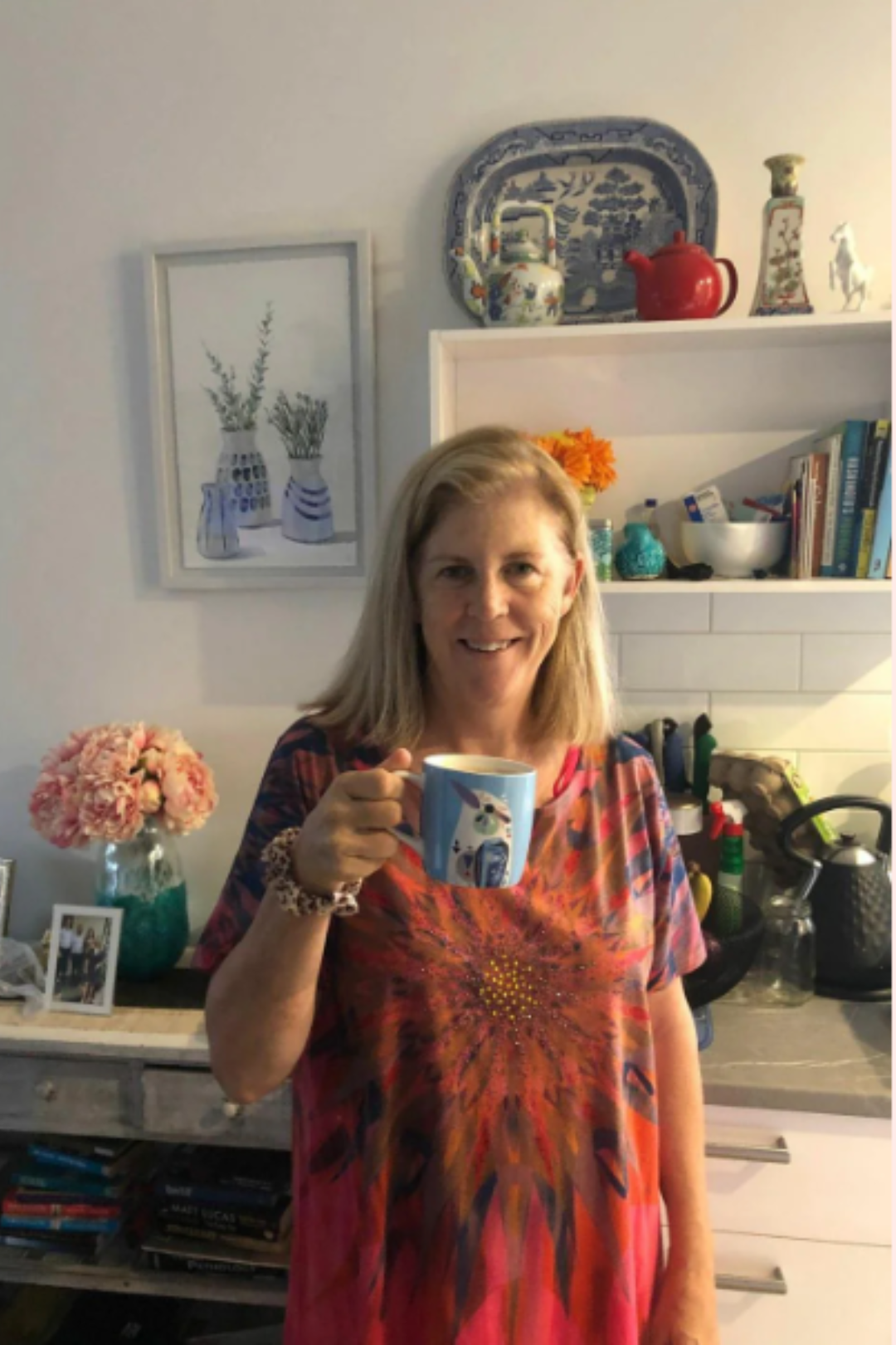 Woman holding a mug in a kitchen with decorative items on shelves.