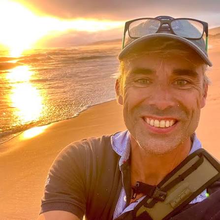 A smiling man takes a selfie on a beach at sunset, with the ocean in the background.