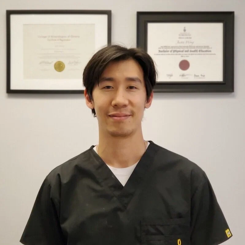 Person in black scrubs standing in front of framed certificates on a wall.