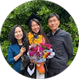 A young female graduate in a cap and gown smiles with her parents while holding a large flower bouquet.
