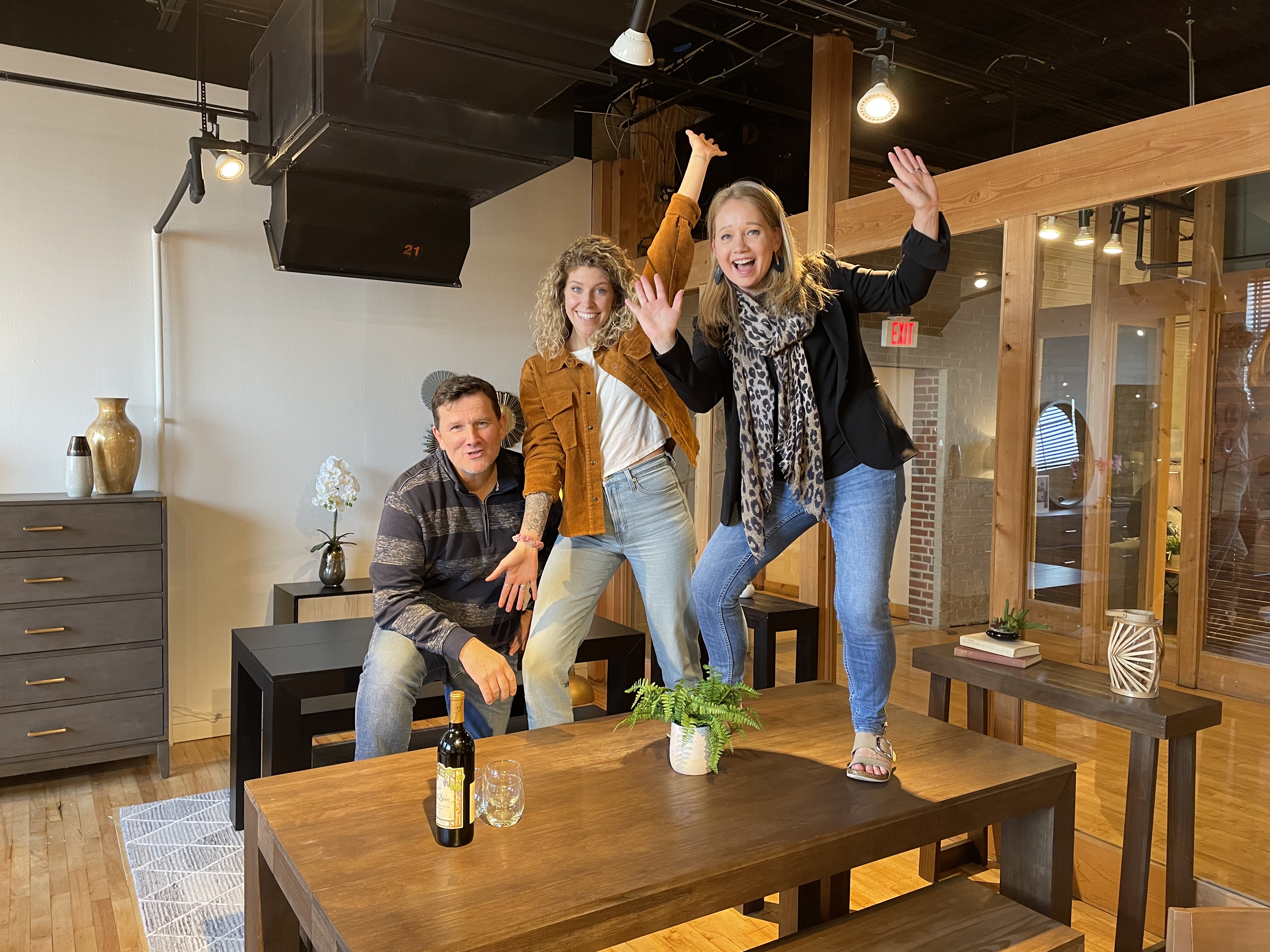 Three people posing energetically in a modern office setting with wooden furniture.