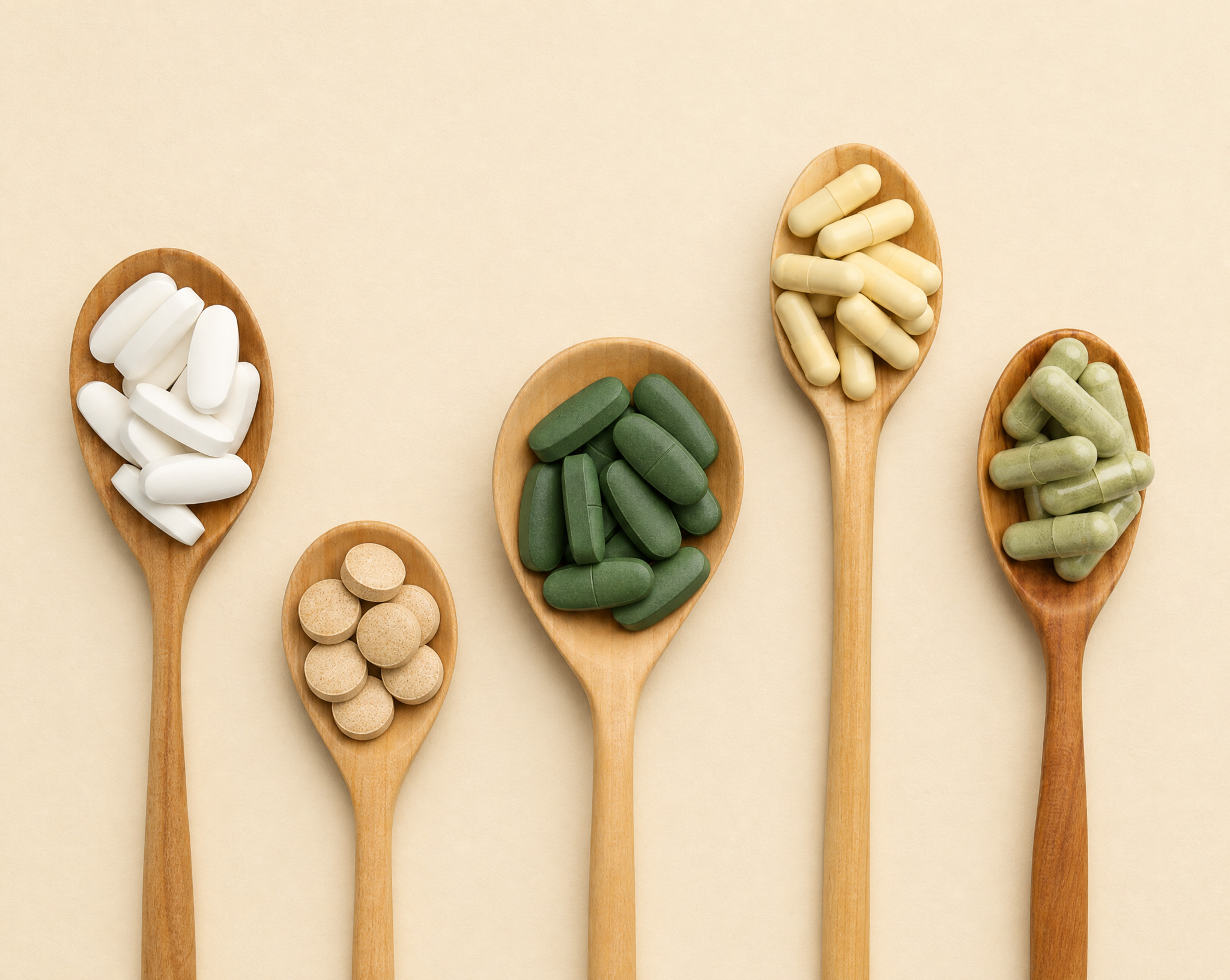 An overhead shot of five wooden spoons, each containing different vitamin pills, tablets, and capsules.