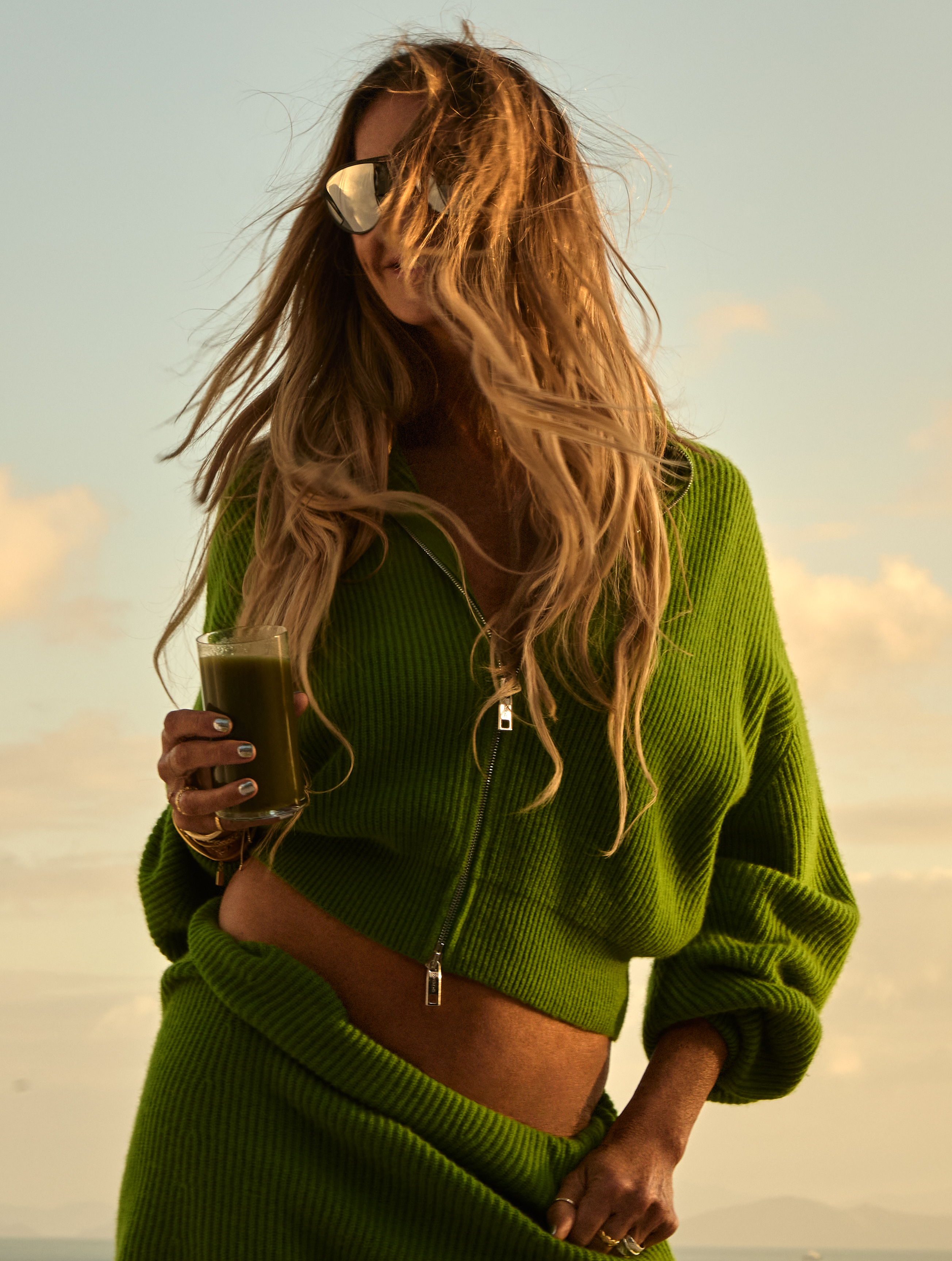 A woman in a green outfit and sunglasses holds a glass of green juice, her hair blowing.