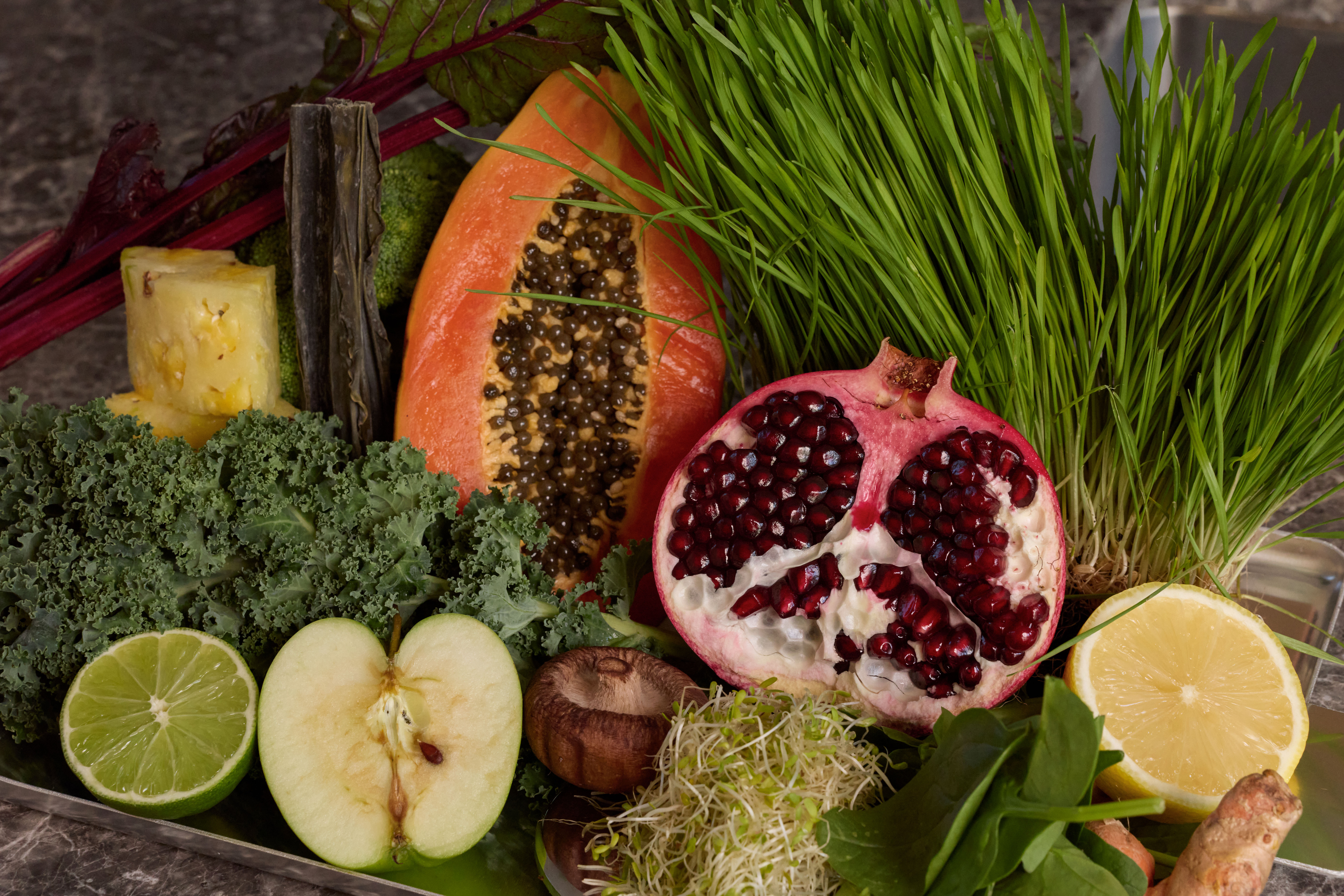 A colorful assortment of fresh fruits and vegetables, including pomegranate, papaya, wheatgrass, kale, and citrus.
