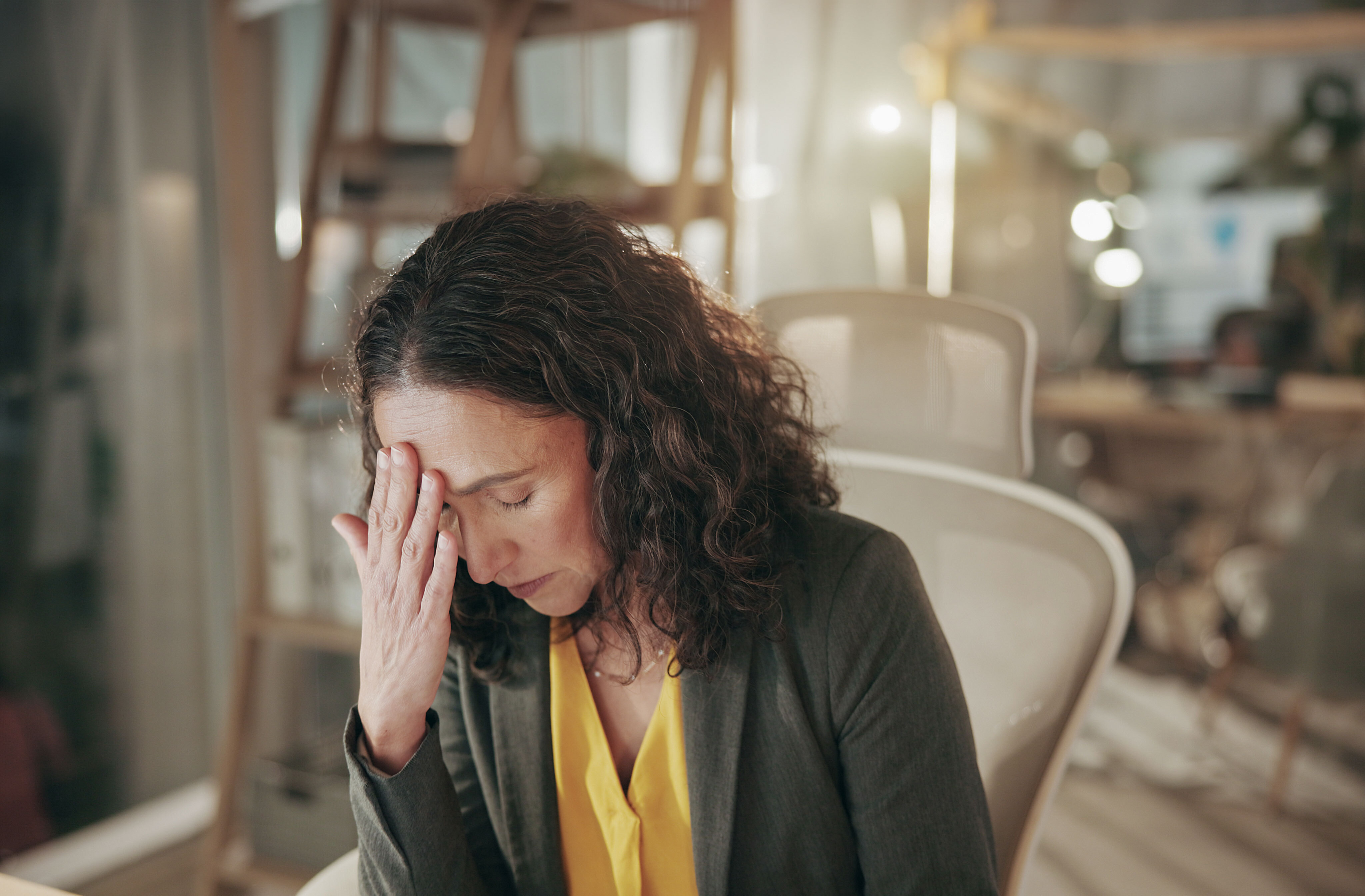 A woman with her eyes closed sits in an office, holding her hand to her forehead.