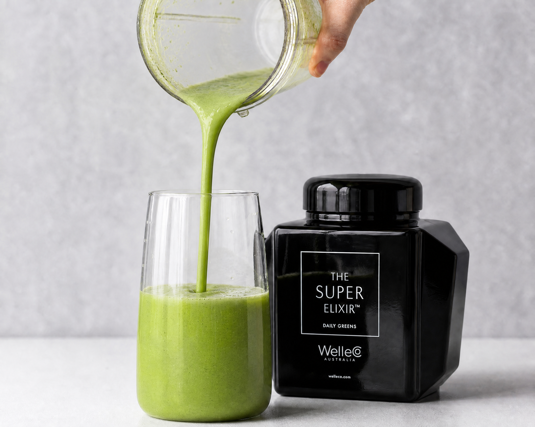 A hand pours a green smoothie from a blender into a glass next to a Super Elixir container.