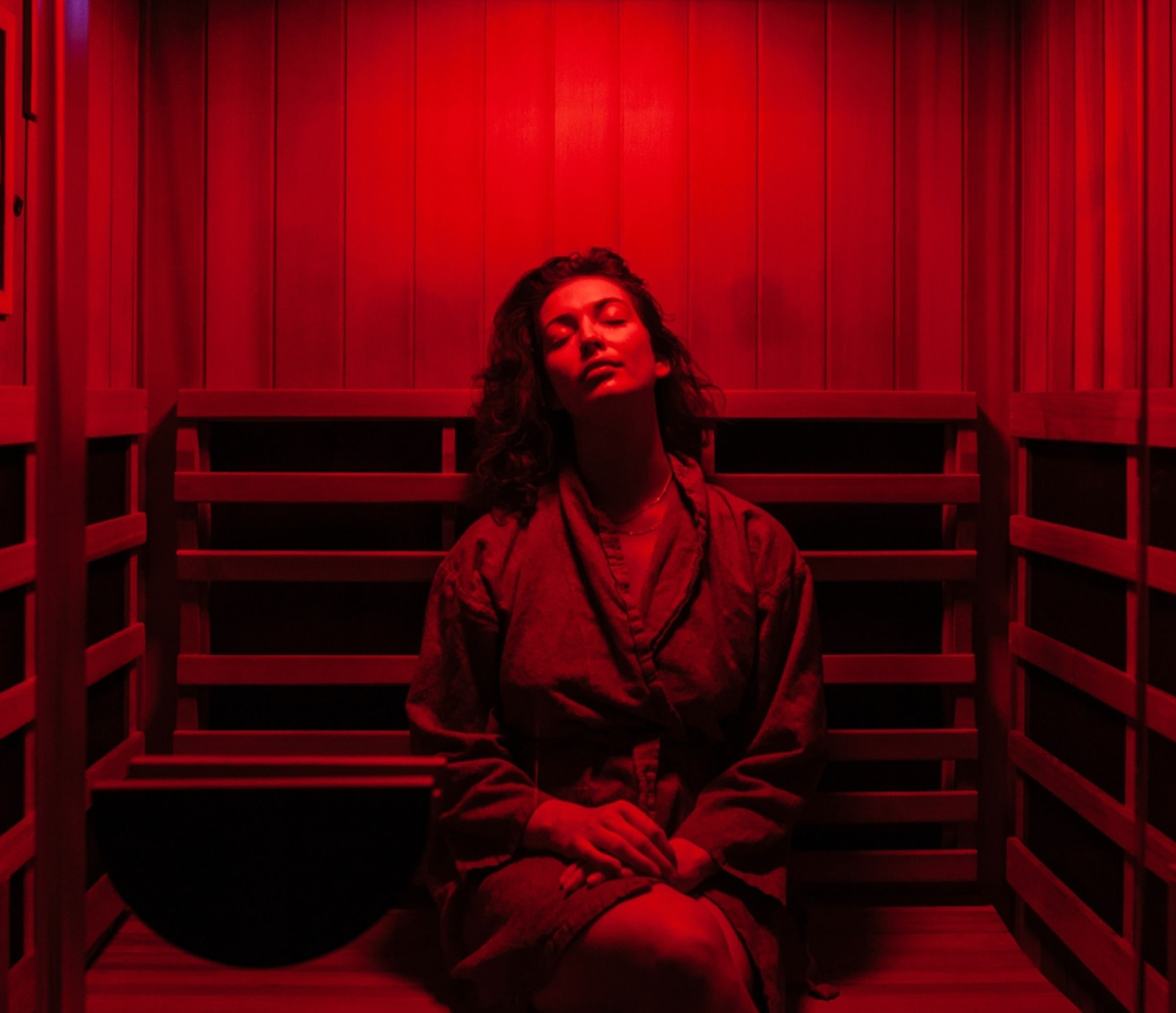 Person sitting in a red-lit sauna, eyes closed, wearing a robe.