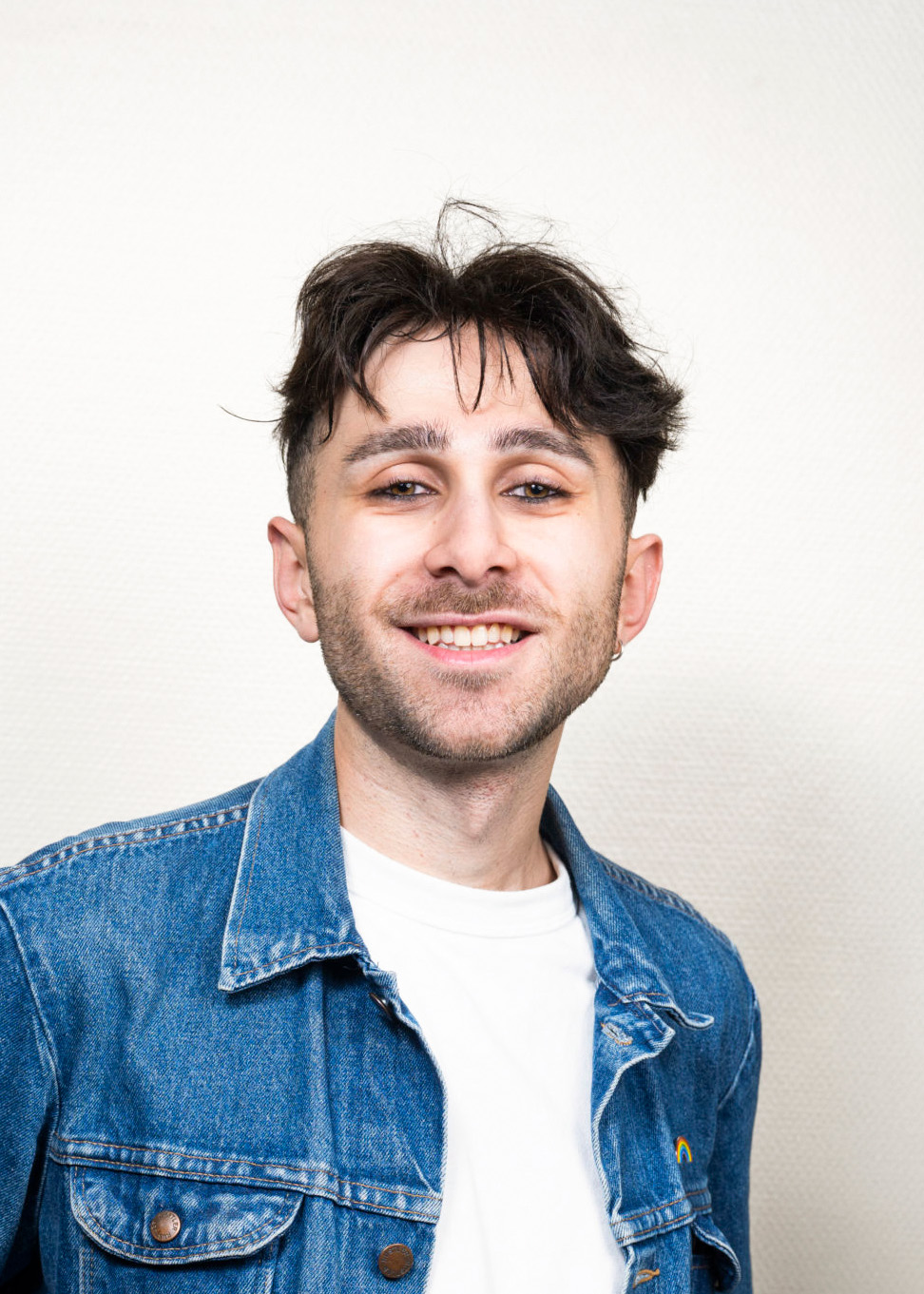 A young man with dark hair in a denim jacket and white t-shirt smiles against a white background.