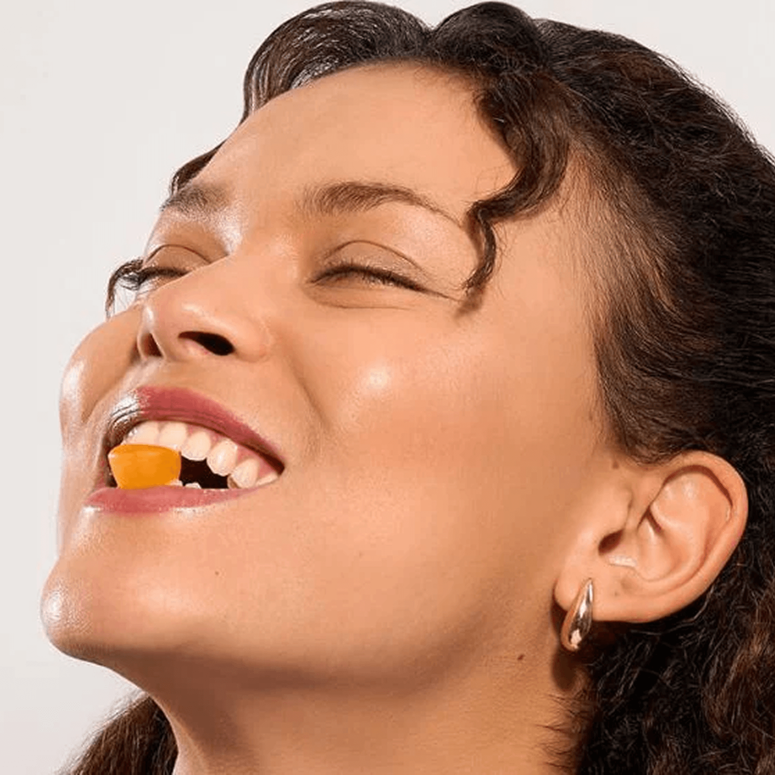 A woman with her eyes closed smiles while holding an orange gummy between her teeth.
