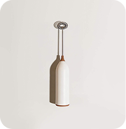 A handheld milk frother with a white handle and metal whisk, standing against a light-colored wall.