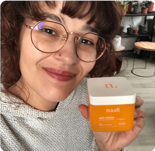 Person smiling, holding a jar labeled 'naali Anti-Stress' in a room with plants.