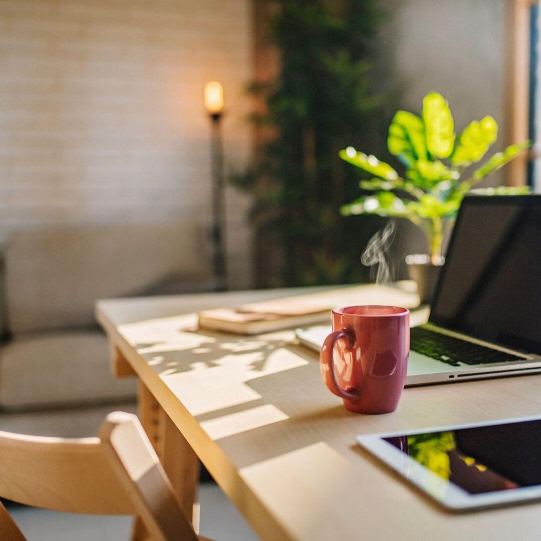 Cozy workspace with a laptop, tablet, and steaming mug on a wooden table.