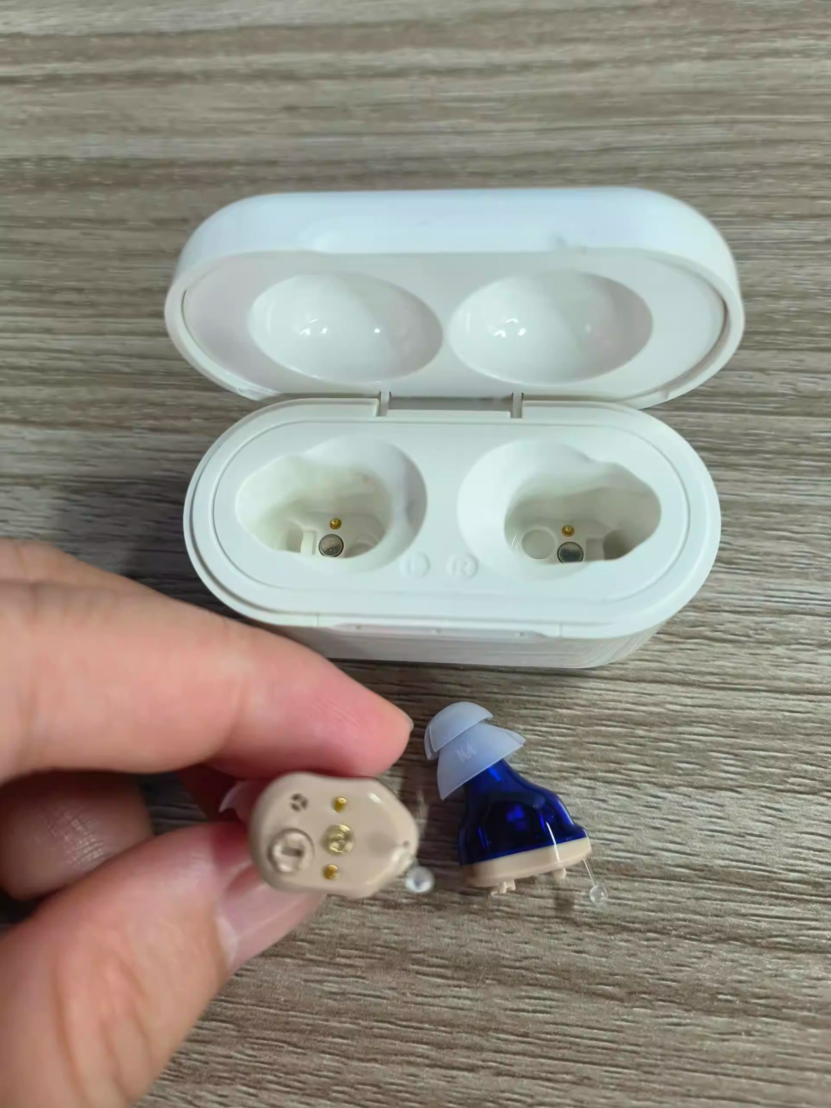 A hand holding a small hearing aid next to another one and their white charging case.
