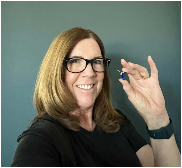 A smiling woman with glasses and brown hair holds up a small blue earring against a gray-blue wall.