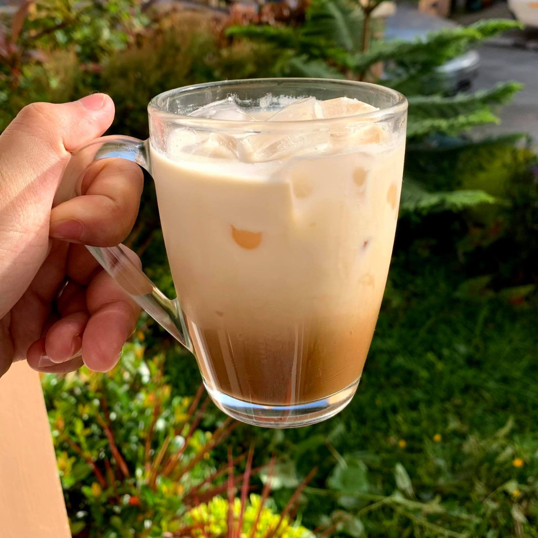 Hand holding a glass mug of iced coffee with milk outdoors.