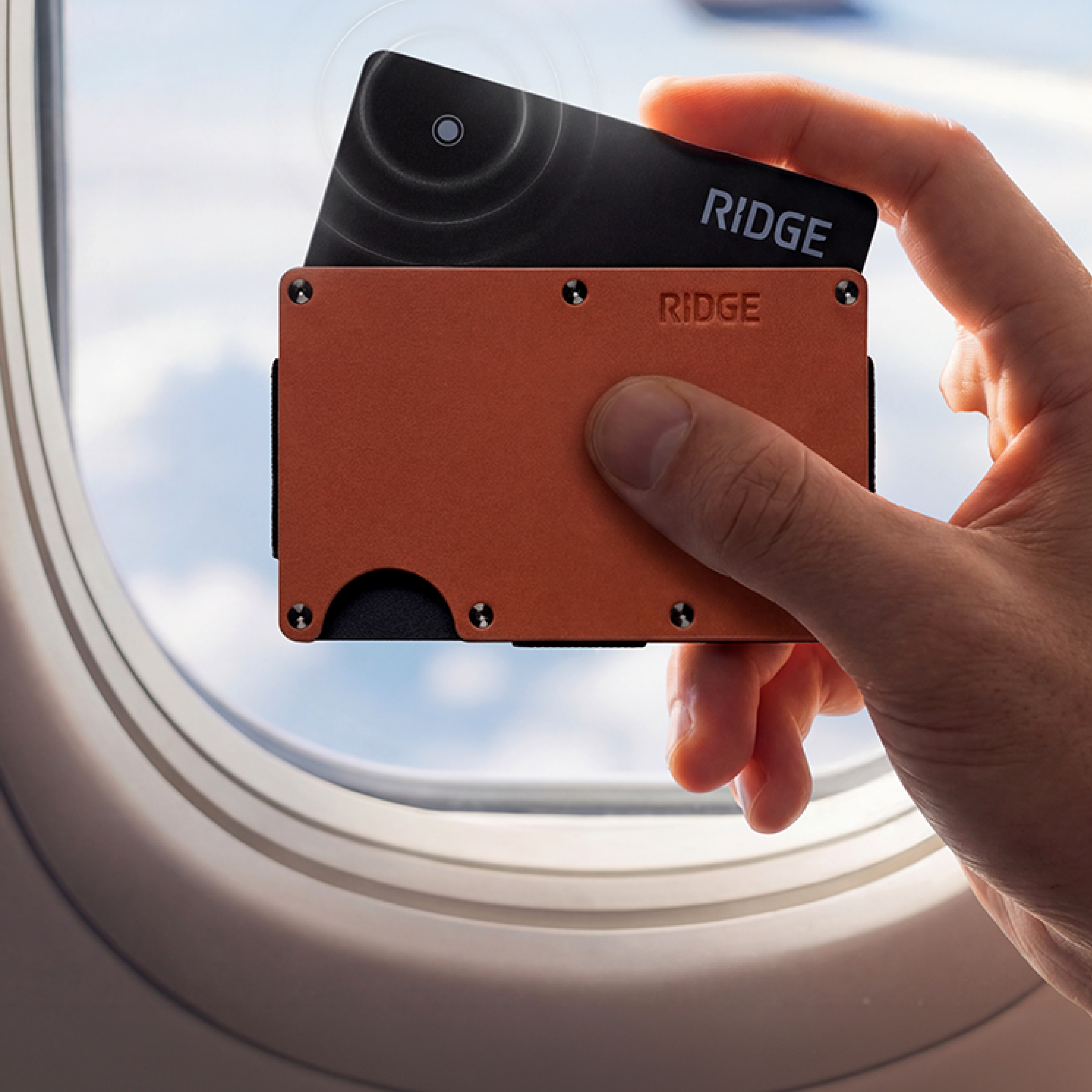 A hand holding an orange Ridge wallet and a black card by an airplane window.