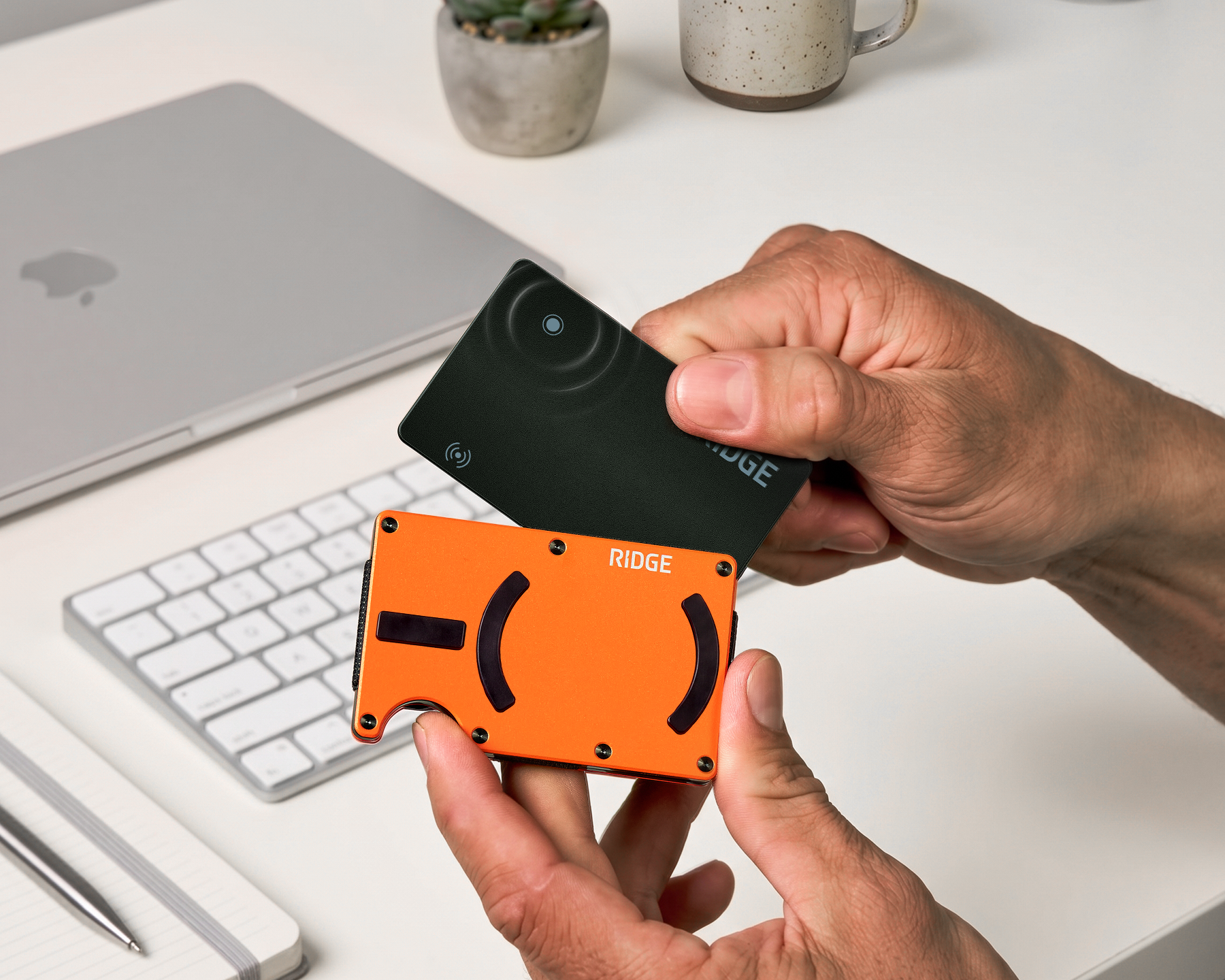 A person inserts a black card into an orange Ridge wallet over a desk with a laptop.