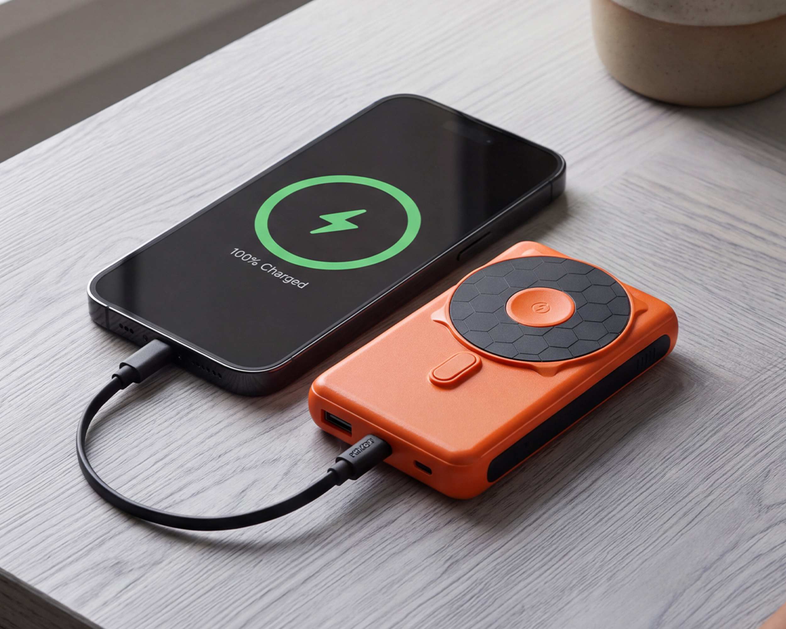 An orange portable power bank charging a smartphone that is at 100% battery on a wooden table.