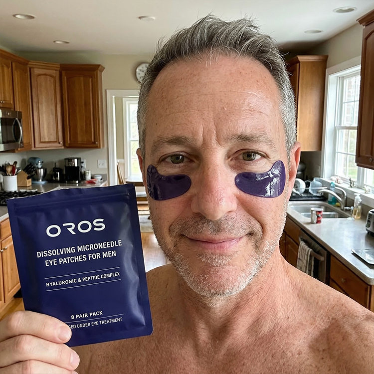 A smiling man with purple patches under his eyes holds up a package of Oros eye patches.
