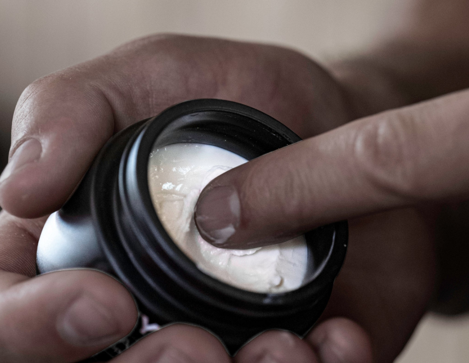 A close-up of a person's finger dipping into a small black jar of white cream.