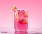 A glass of a pink beverage with strawberries and a watermelon wedge against a solid pink background.