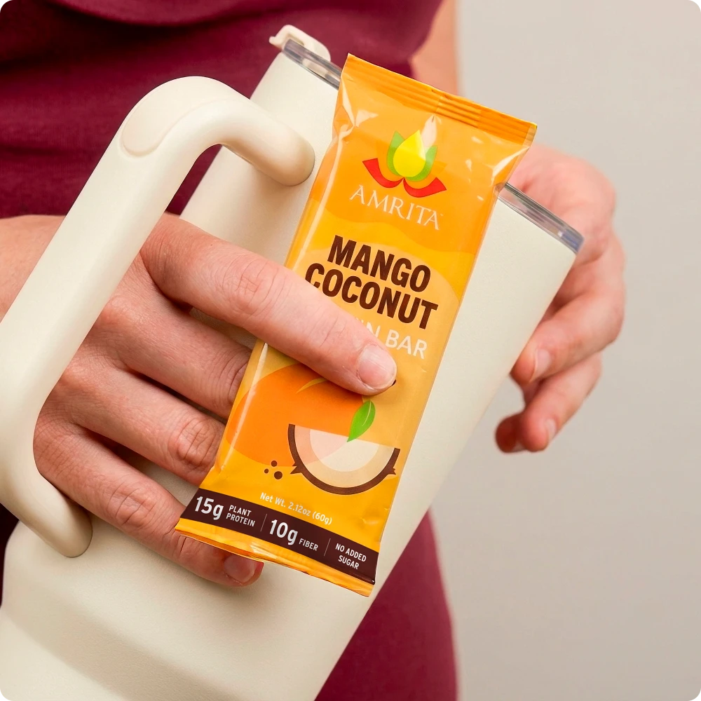 Close-up of a hand holding an Amrita Mango Coconut Protein Bar package against a white tumbler, highlighting 15g of plant protein and 10g of fiber.