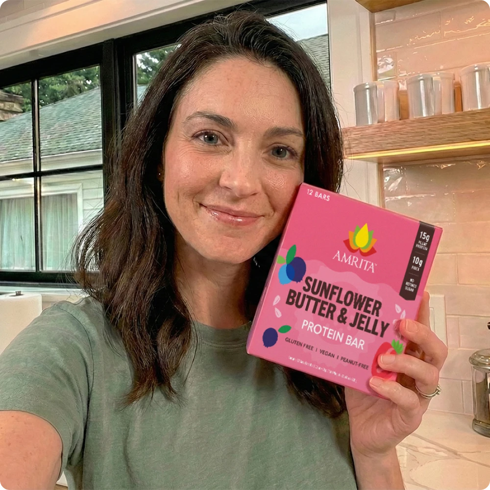 Woman holding a box of Amrita Sunflower Butter & Jelly protein bars in a bright kitchen setting