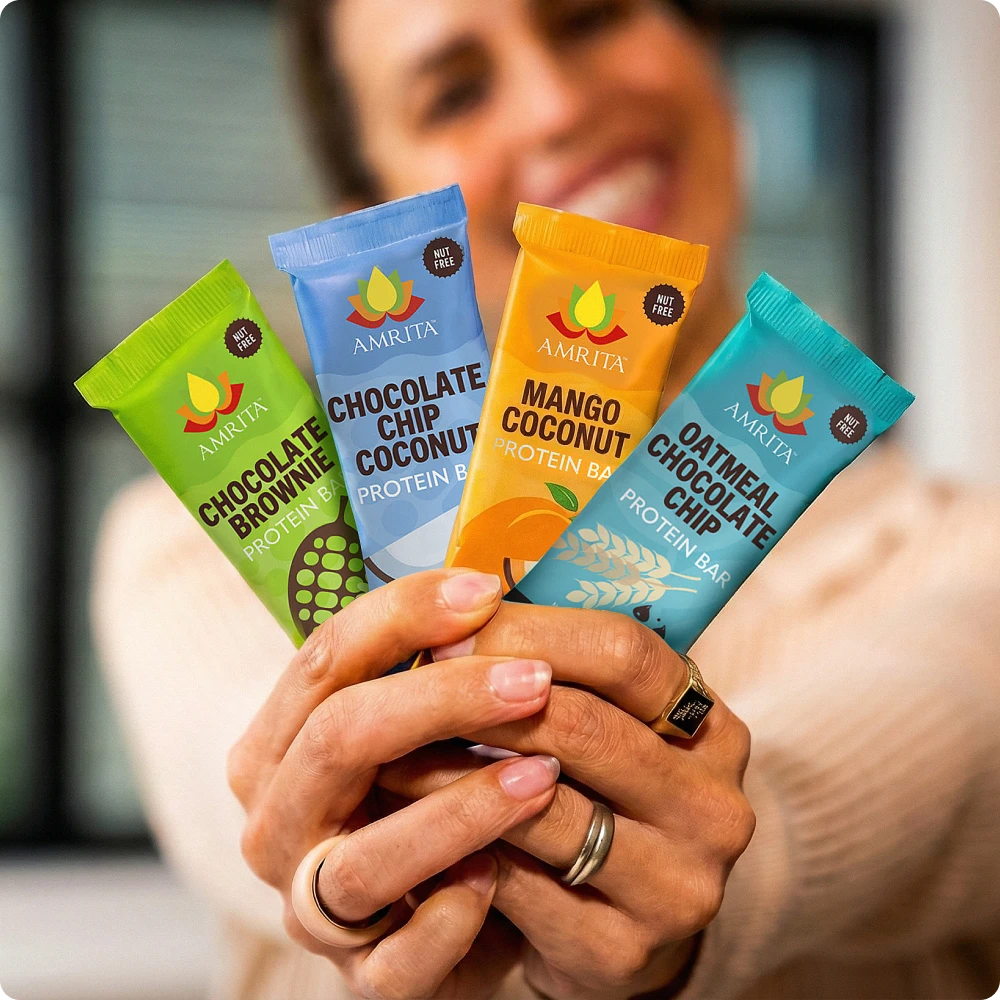 Smiling woman holding a variety of four Amrita Protein Bars fanned out in her hands, showing the Nut Free label.