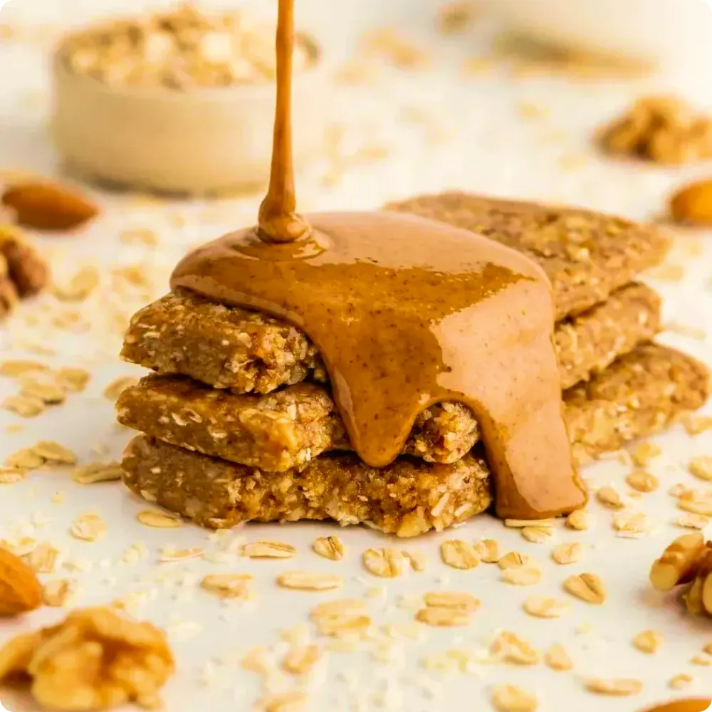 Protein bars stacked with creamy nut butter dripping on top, surrounded by oats and nuts on a light background
