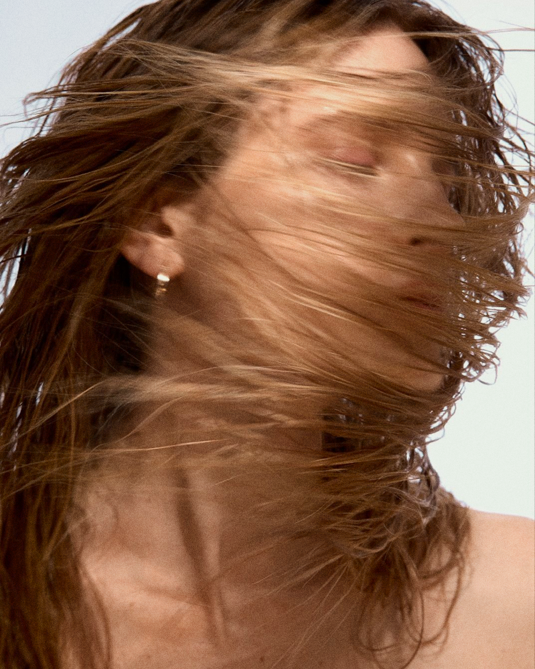 A close-up of a person with eyes closed as their hair blows across their face, revealing an earring.