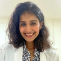 Person smiling, wearing a white coat and checkered shirt, with a blurred background.