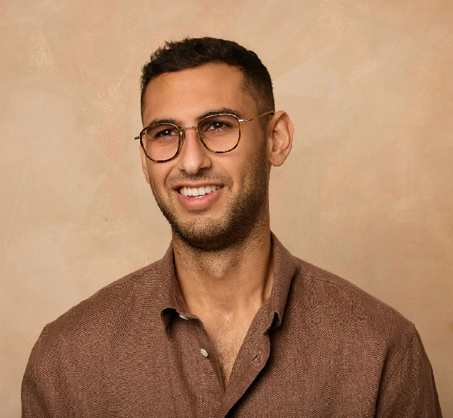 Man with glasses smiling, wearing a brown shirt against a beige background.