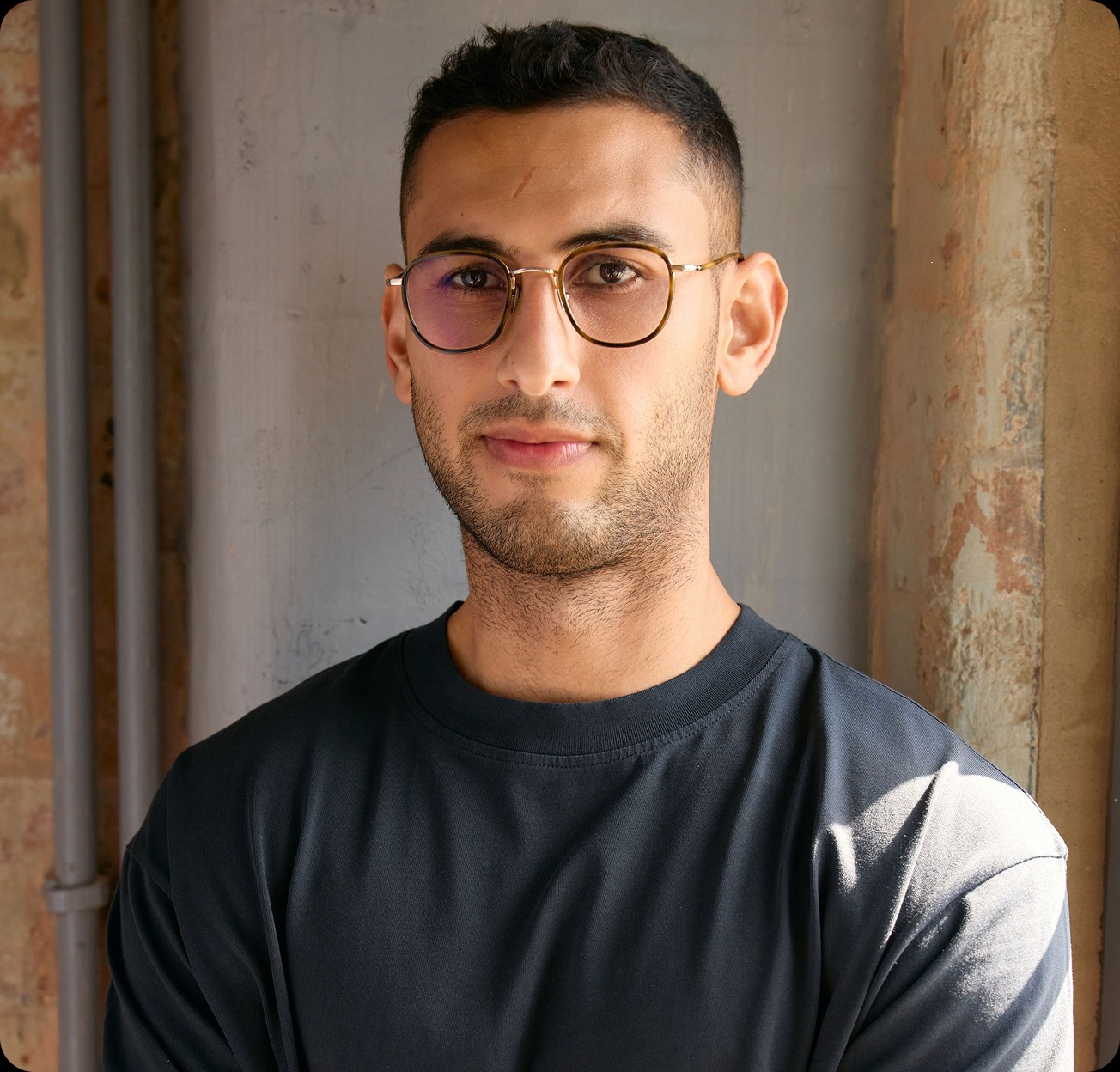 Person wearing glasses and a dark shirt, standing indoors by a textured wall.