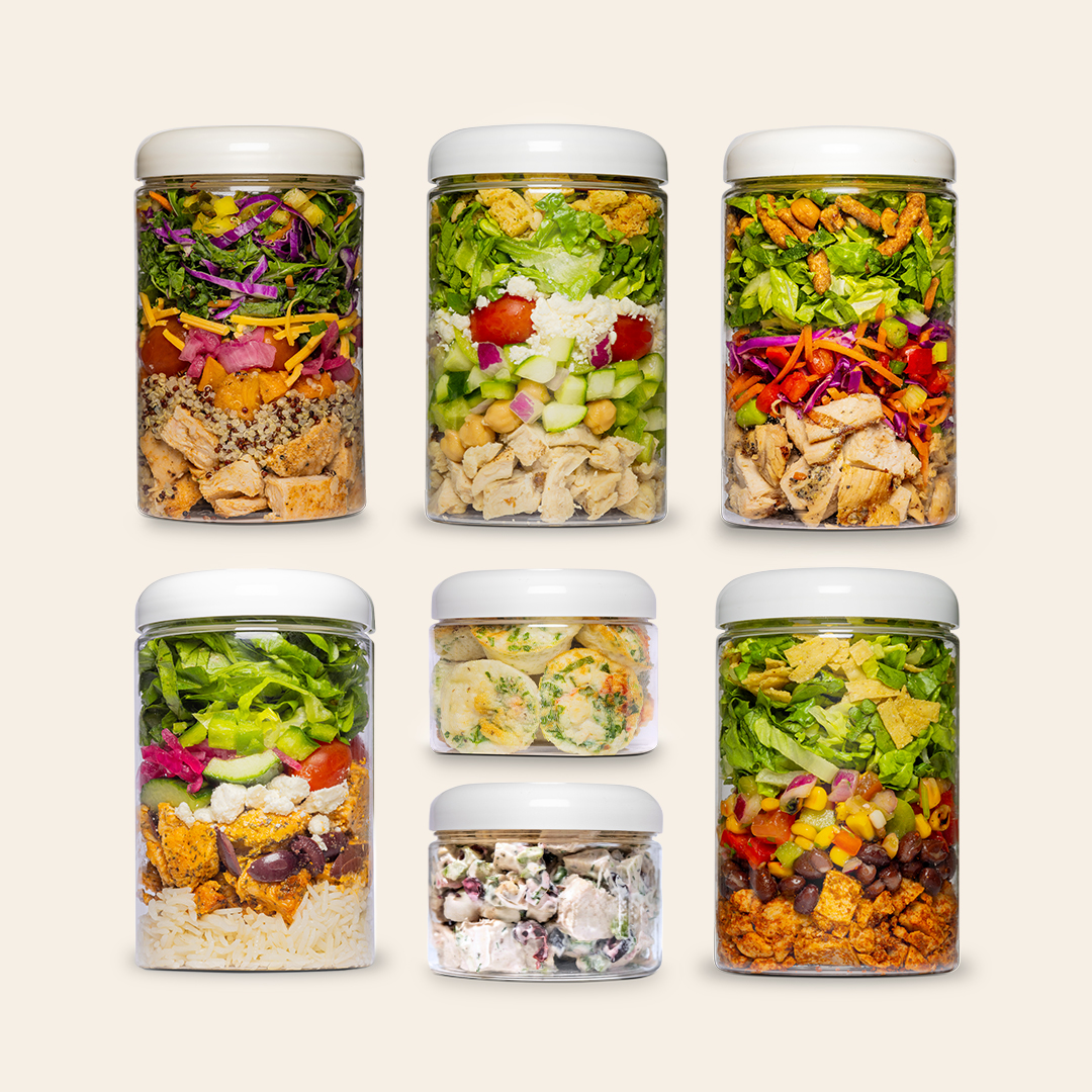 Six clear jars of various sizes filled with colorful, layered salads and other meals.