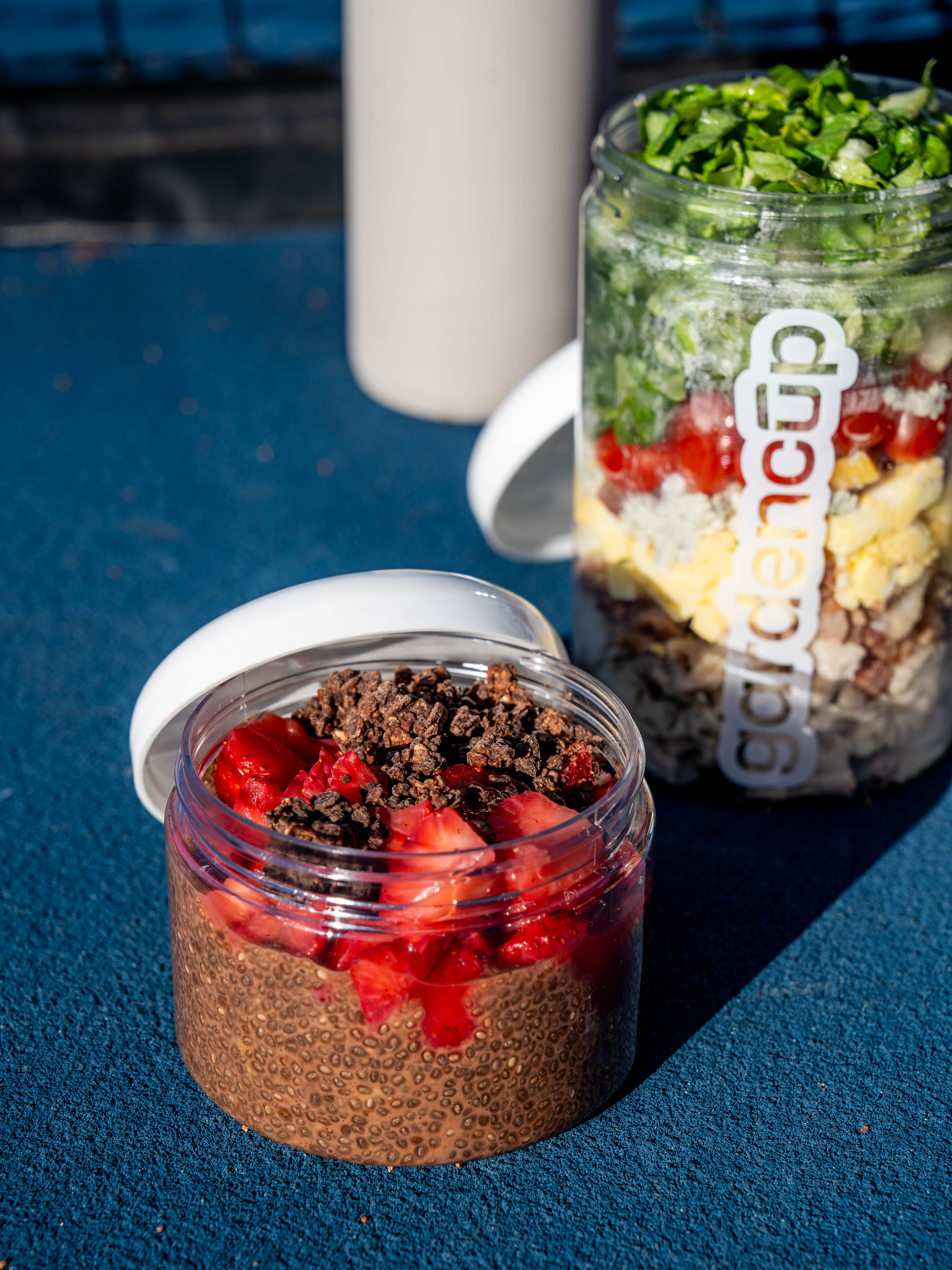 Two jars with chia pudding and salad on a blue surface.