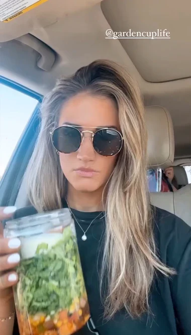A woman with blonde hair and sunglasses sits in a car holding a salad in a jar.